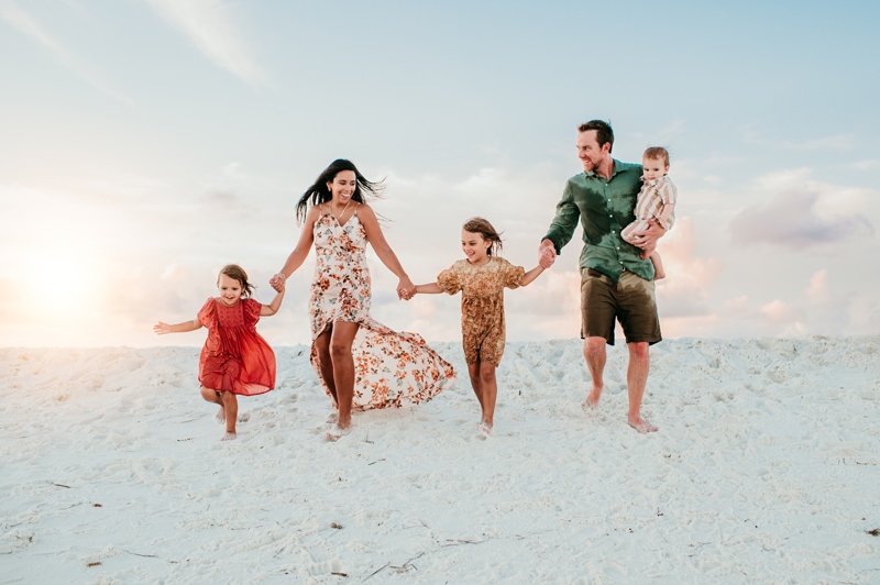 Family Photographer, a mother and father walk on the beach with their children while holding baby